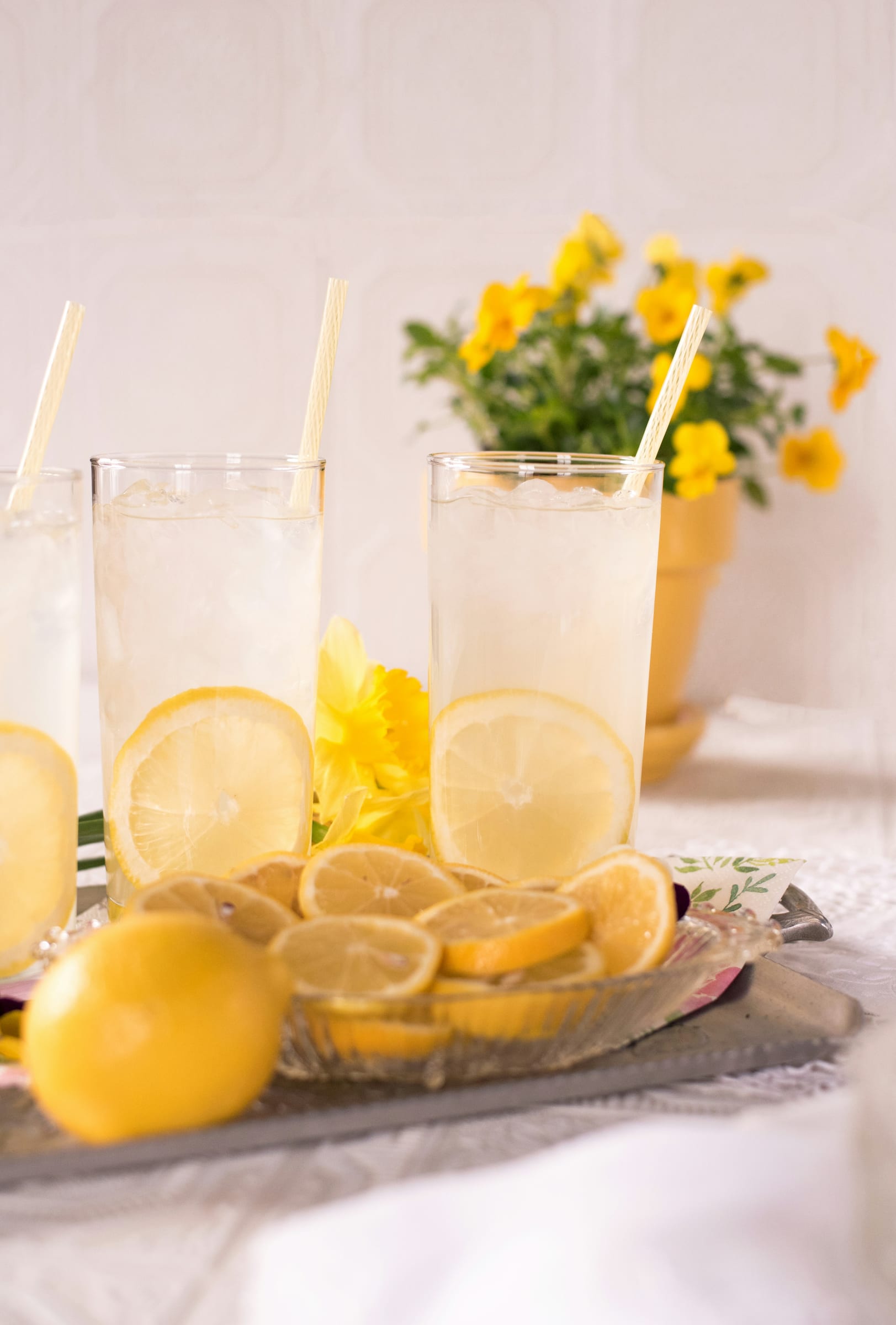 Tall glasses of iced lemonade with lemon slices and paper straws on a tray, with yellow flowers behind