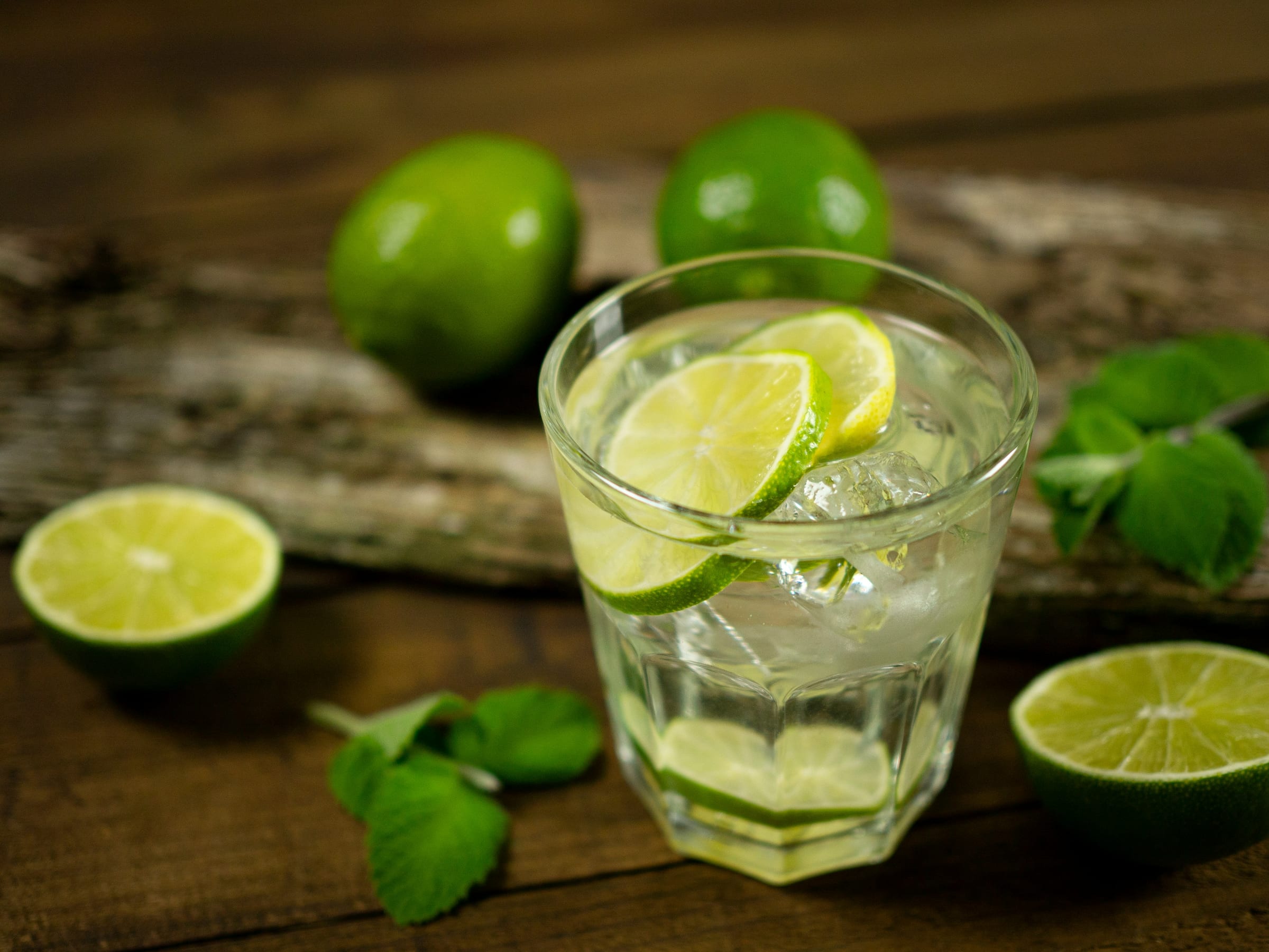 A glass of ice water with lime slices on a rustic wood table, surrounded by whole and halved limes and fresh mint