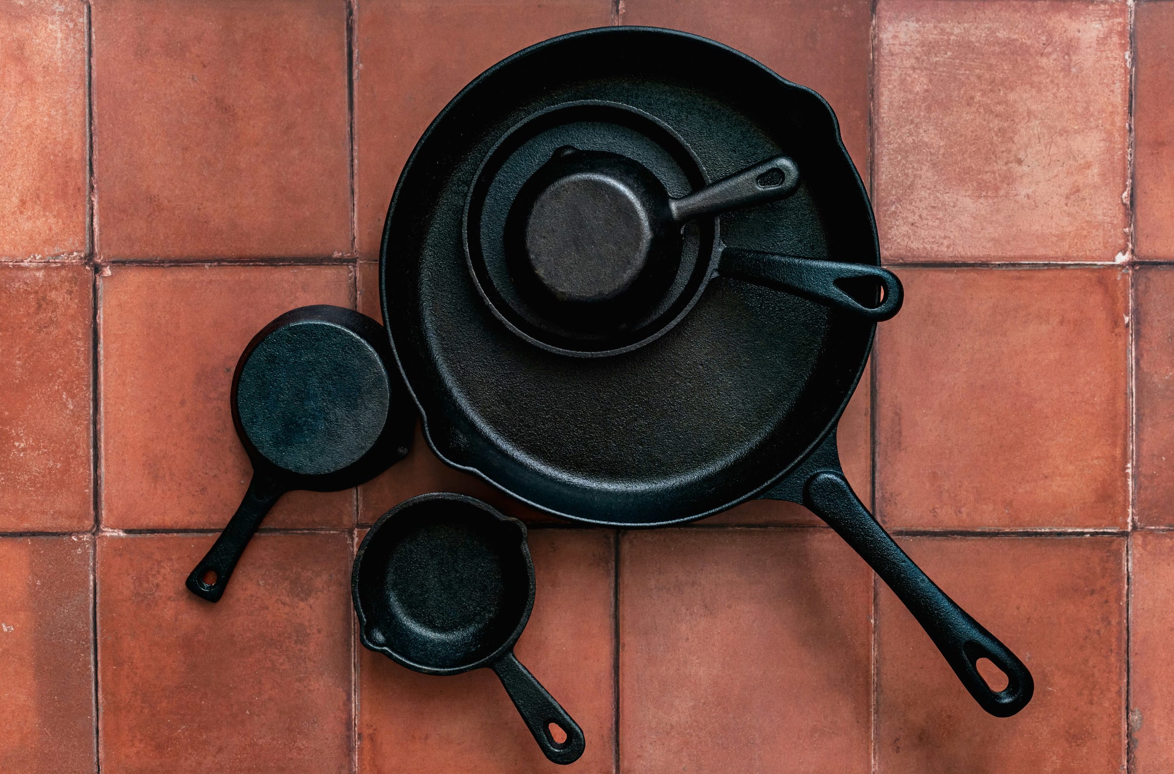 A set of well-seasoned cast iron skillets arranged on warm terracotta tile