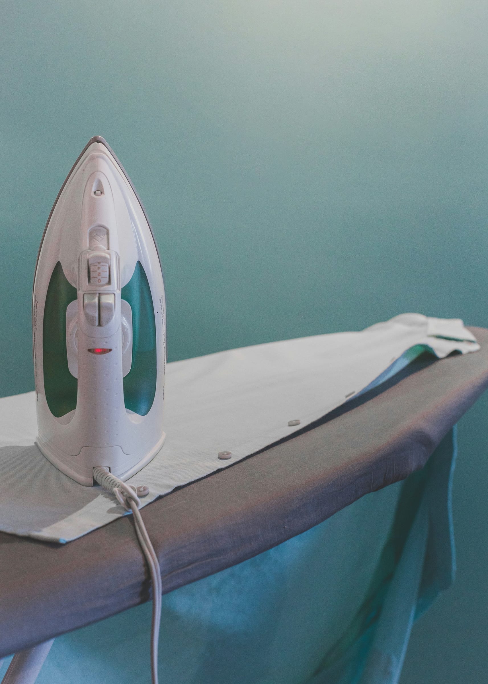 A white steam iron standing upright on an ironing board against a teal wall