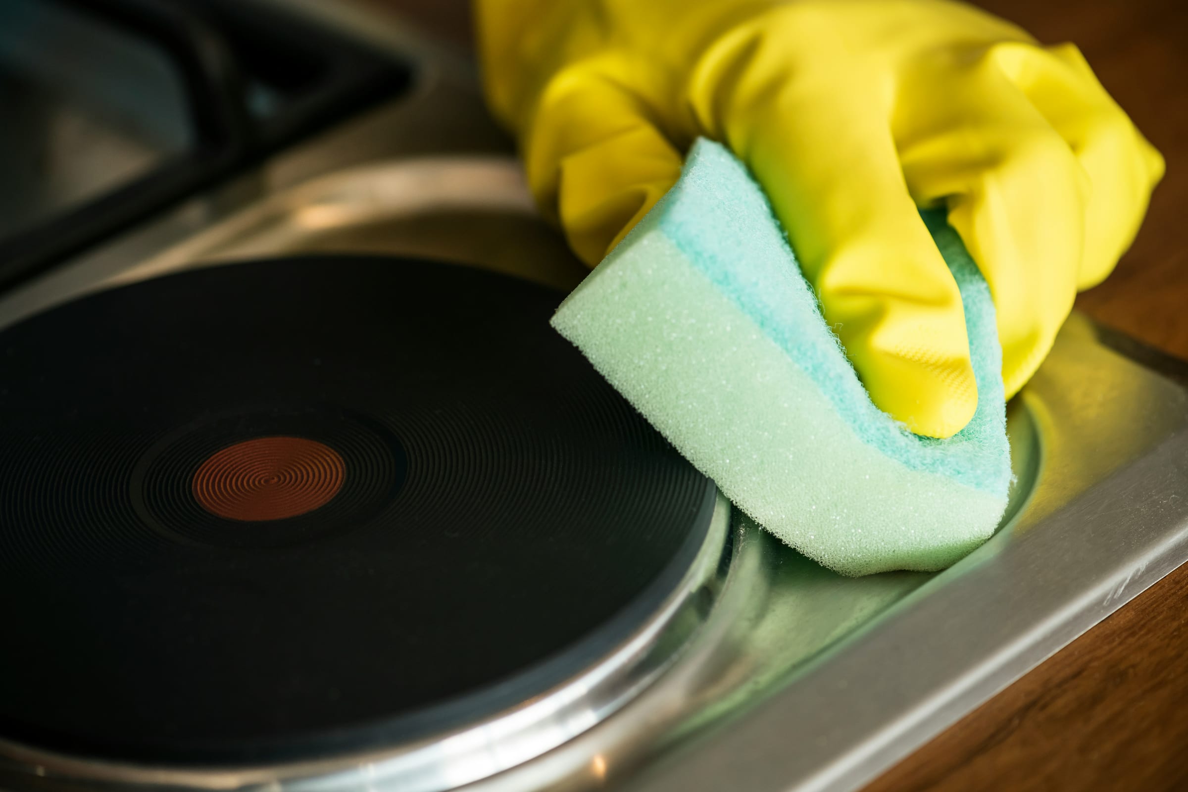 A hand in a yellow rubber glove scrubbing a black stovetop burner with a sponge