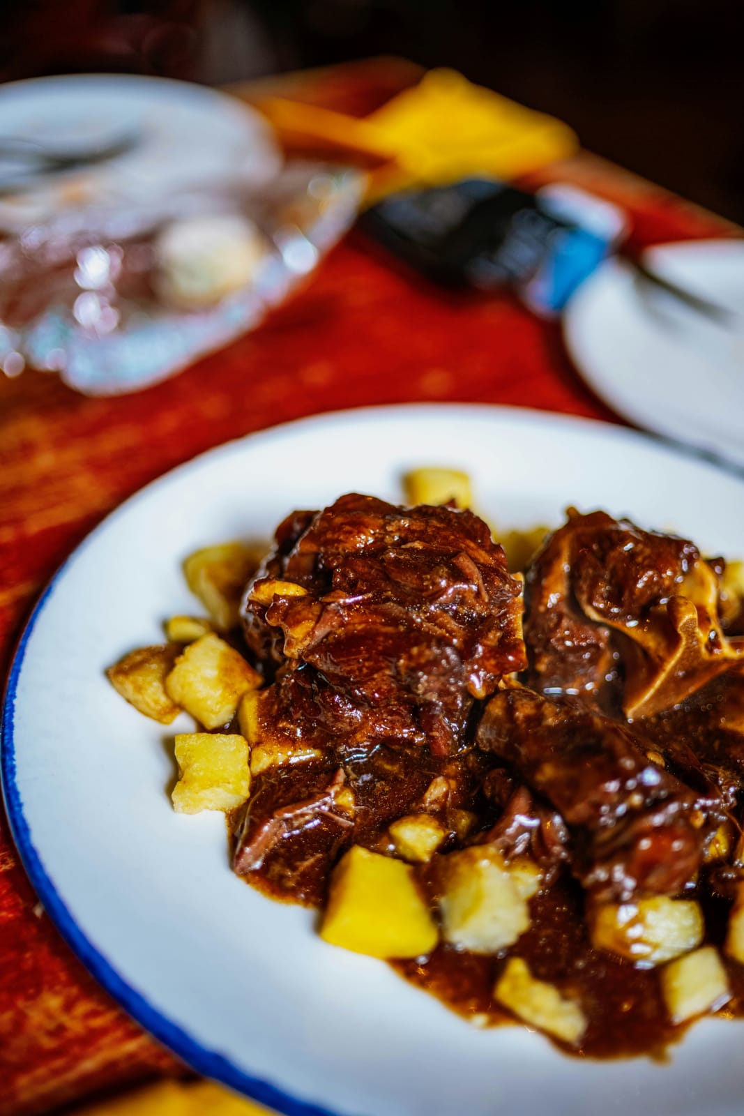 Braised oxtails glazed in a glossy brown gravy over golden cubes of potato on a blue-rimmed white plate, set on a red tablecloth