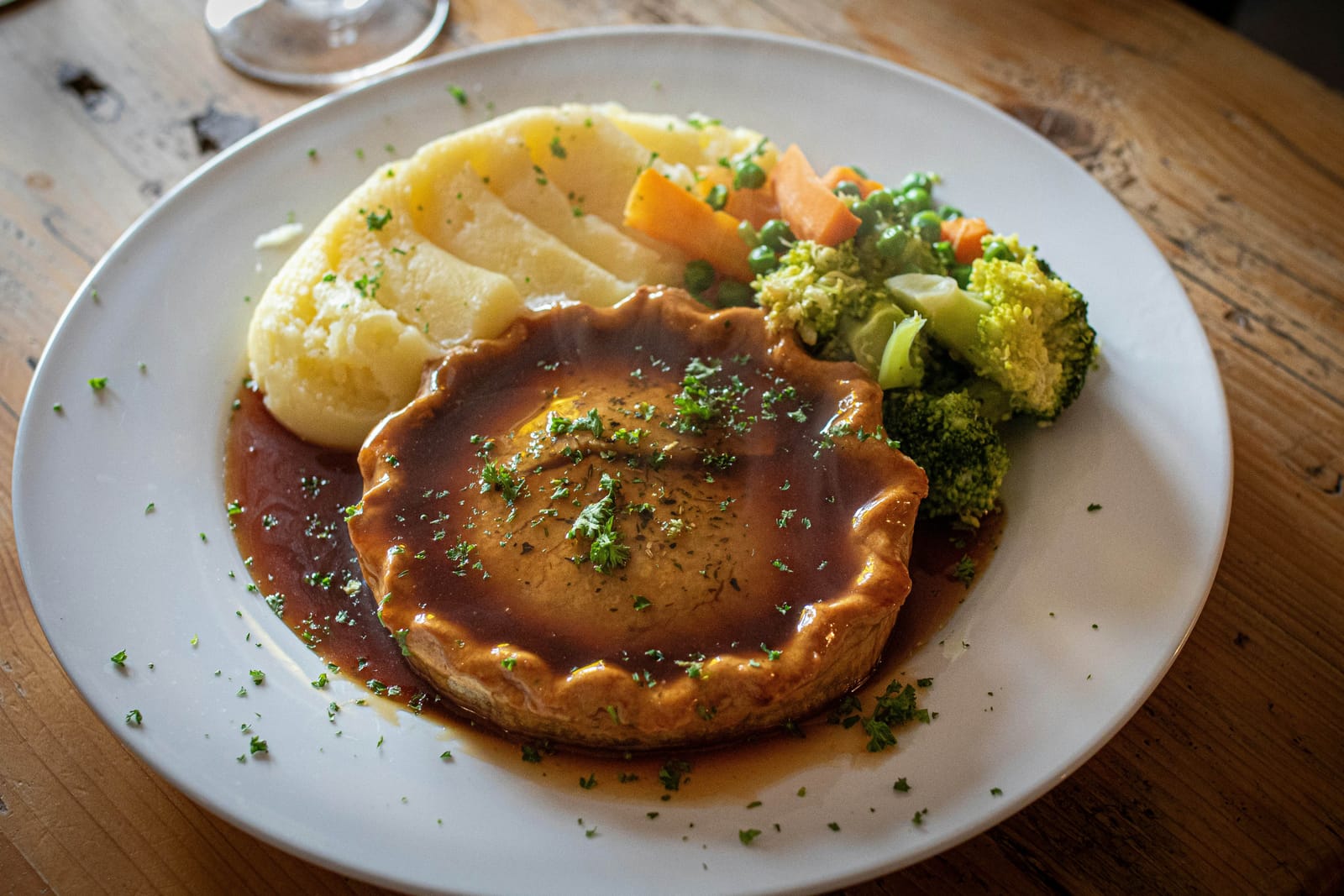 A golden pot pie smothered in brown gravy and sprinkled with parsley, served with mashed potatoes, peas, carrots and broccoli on a white plate