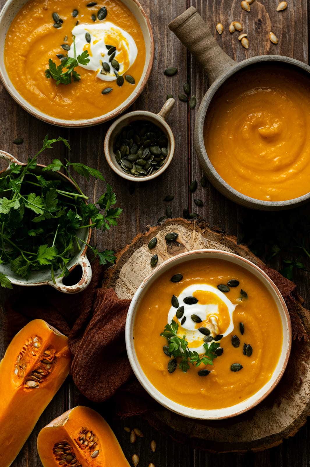 Overhead view of bowls of butternut squash soup swirled with sour cream, topped with pumpkin seeds and parsley, beside halved butternut squash on a dark wood table