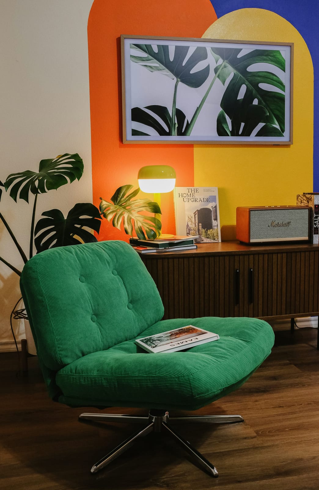 A vibrant living space with an emerald-green corduroy chair against a wall painted in bold orange, yellow, and cobalt-blue arches, a framed monstera-leaf print, a mid-century credenza, a yellow lamp, and lush plants