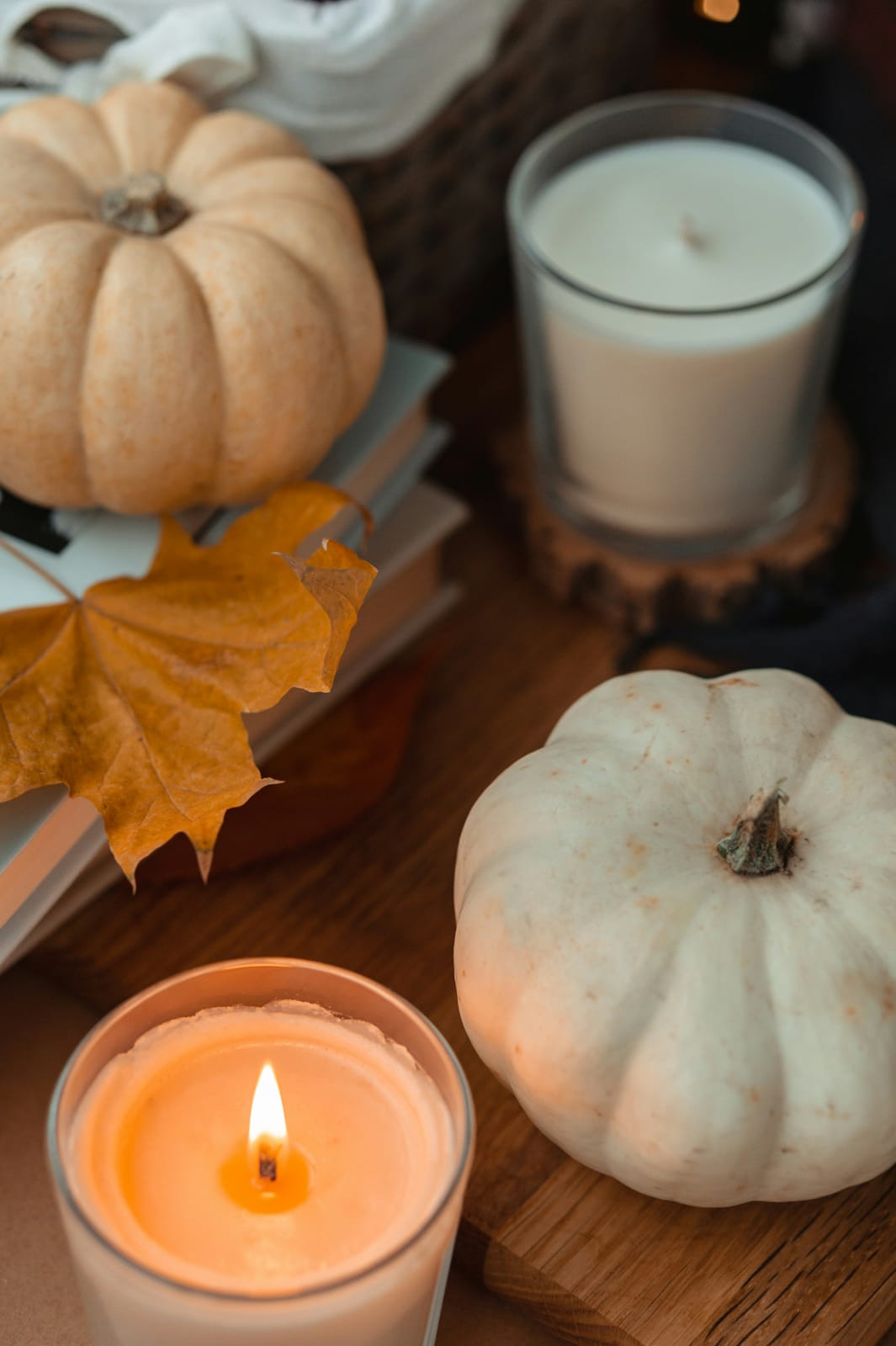 A cozy neutral autumn flat-lay of cream and white pumpkins, white candles with one lit amber flame, a golden maple leaf, and stacked books on a wood surface