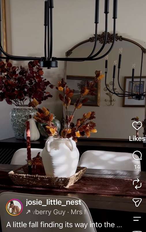 Social post by josie_little_nest: a fall dining table styled with a white ceramic vase of burnt-orange and burgundy autumn foliage on a woven runner, beneath a black candelabra chandelier, captioned “A little fall finding its way into the...”