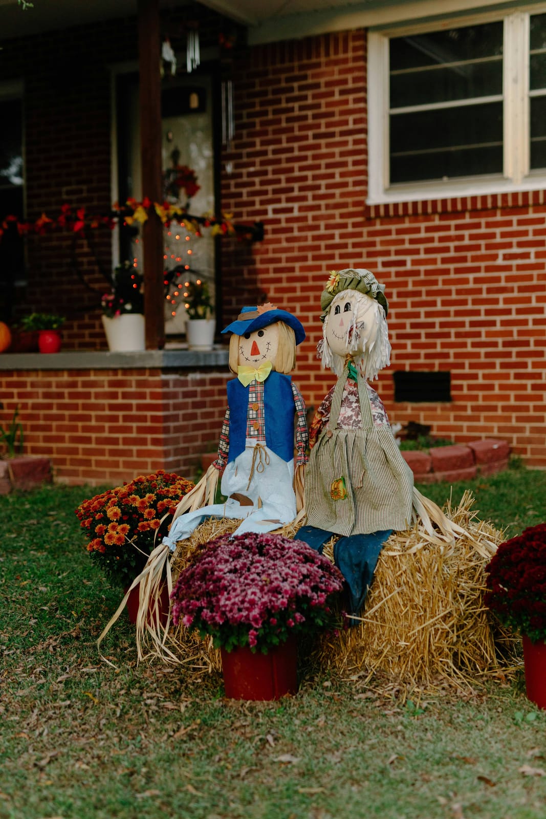 Two burlap scarecrow dolls on a hay bale in a front yard, framed by pots of orange and burgundy mums in front of a red-brick house