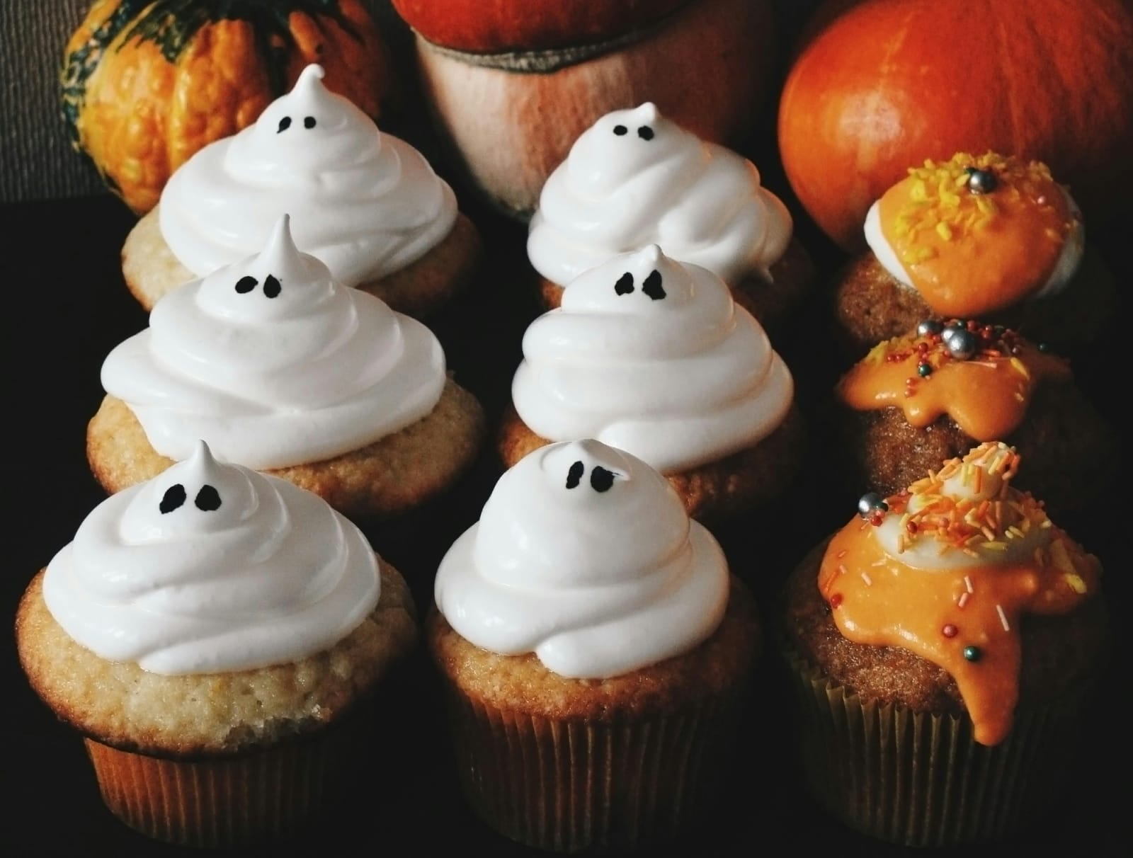 Halloween cupcakes with white meringue ghost toppings and black candy eyes beside orange-frosted cupcakes with fall sprinkles, set against orange pumpkins and gourds