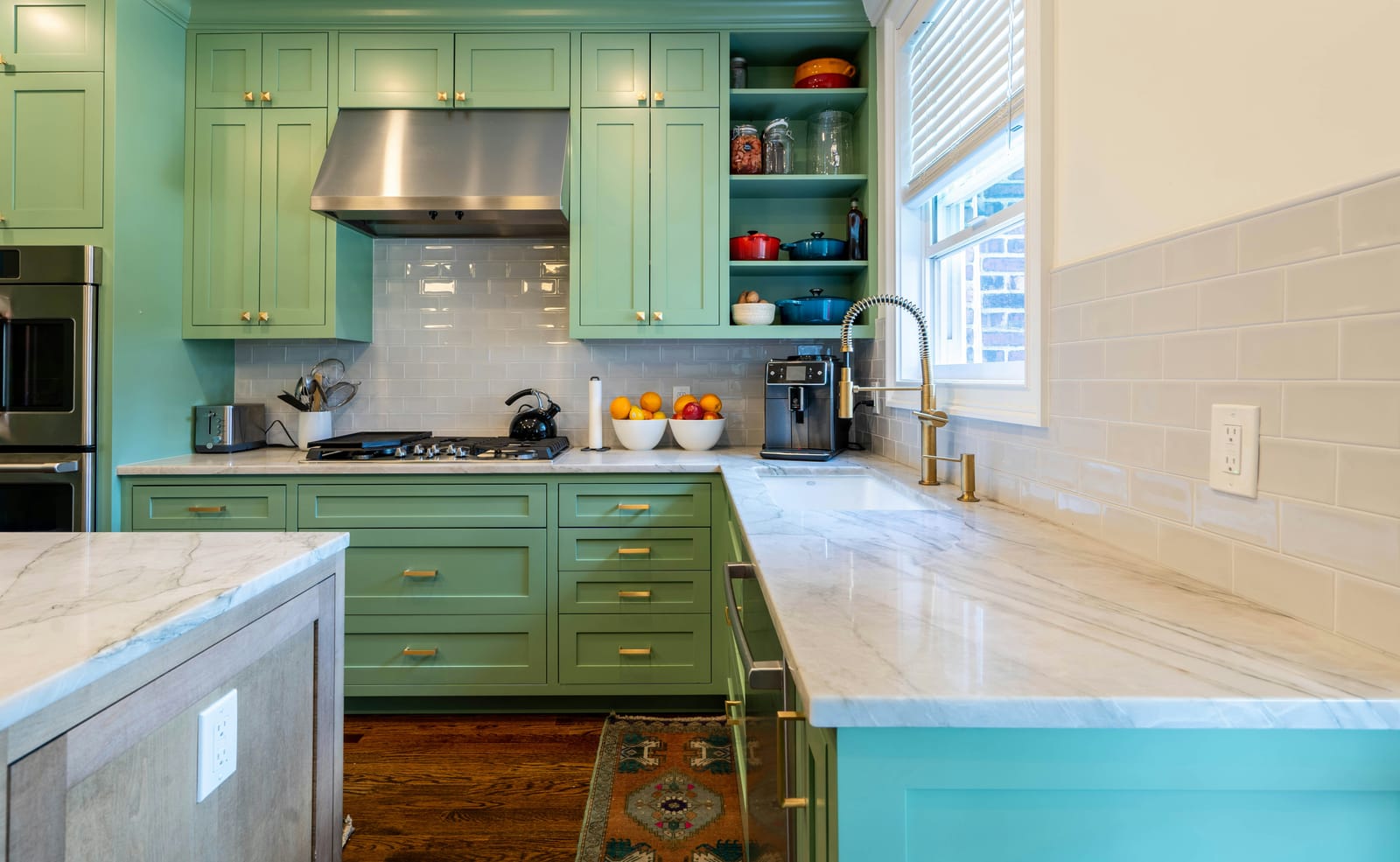 A color-rich kitchen with sage-green shaker cabinets, a teal island, white marble counters, subway-tile backsplash, open shelving with red and blue dutch ovens, and bowls of oranges