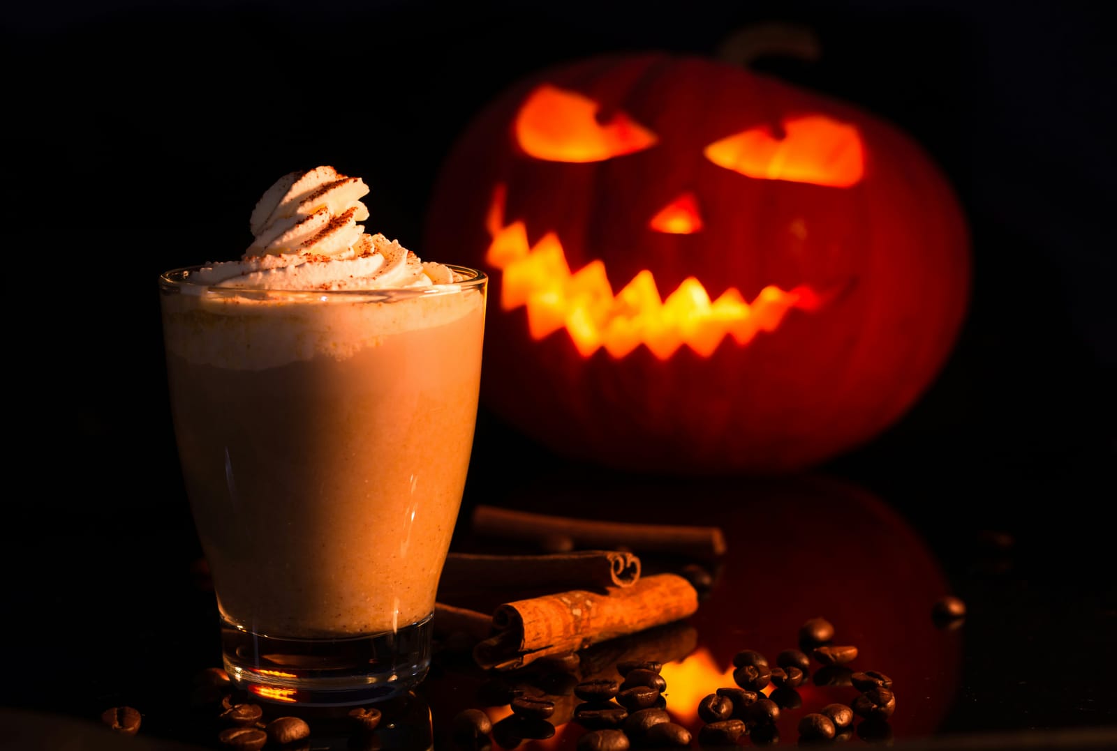 A pumpkin spice latte topped with whipped cream and a dusting of cinnamon in a glass, beside cinnamon sticks and coffee beans, with a glowing carved jack-o’-lantern in the dark background