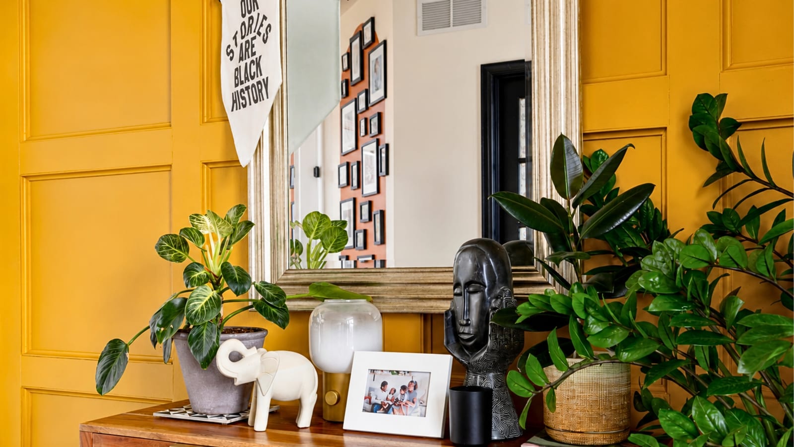 A mustard-yellow paneled entryway with a wood console table holding houseplants, a carved black African stone bust, a white ceramic elephant, and a framed family photo, with an “Our Stories Are Black History” pennant hanging by the mirror