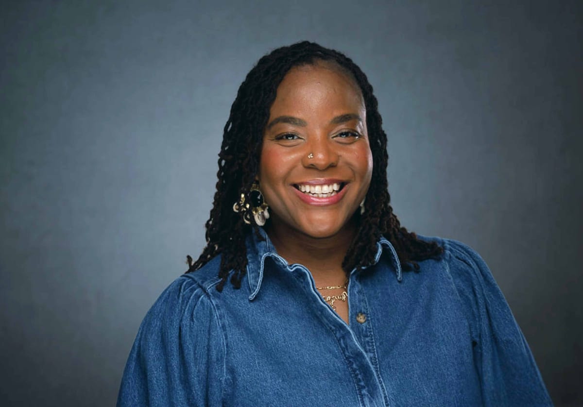 Portrait of interior designer Jazmyn Miller-Gibbs, a smiling Black woman with long locs and gold statement earrings wearing a denim shirt against a soft gray backdrop