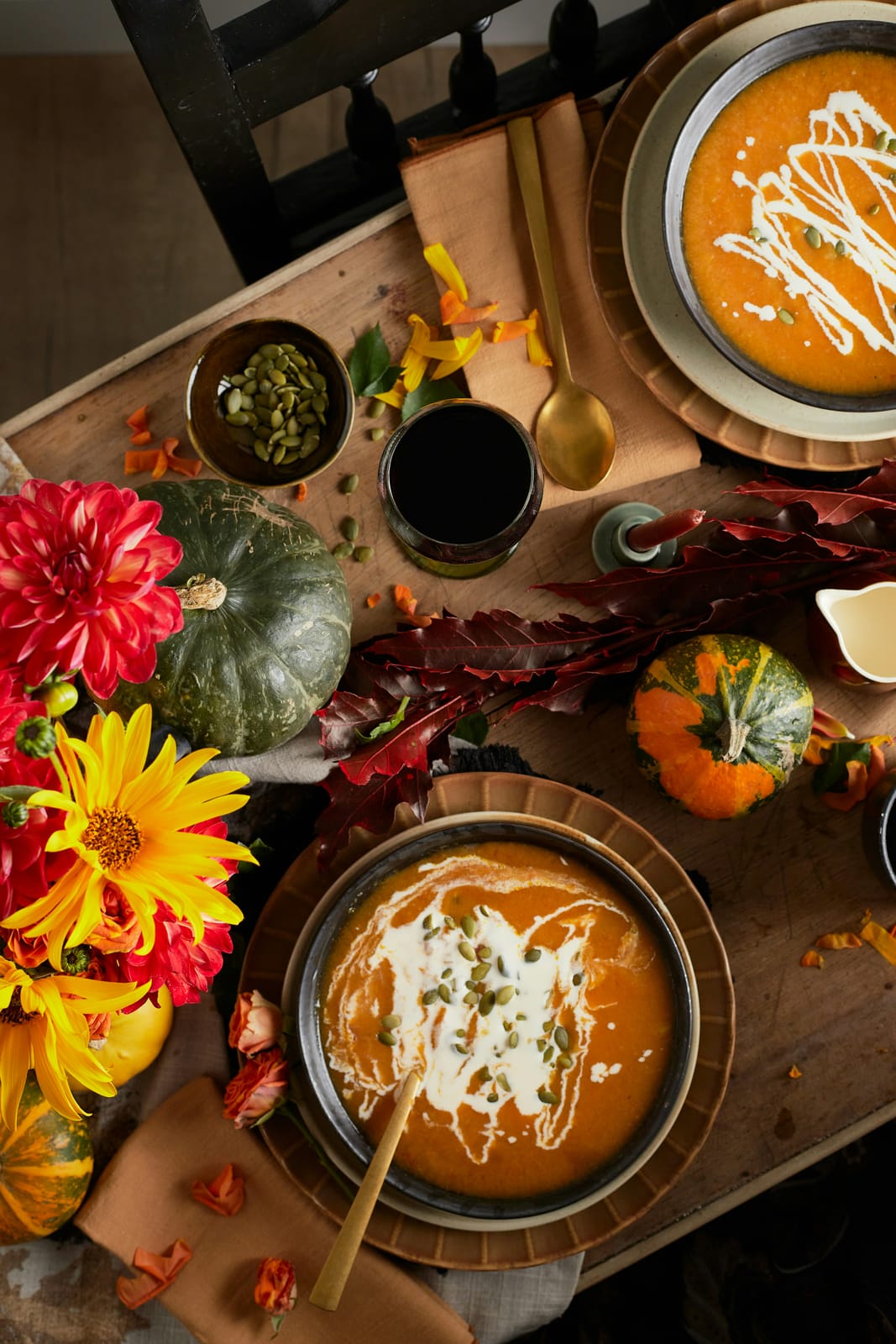 An overhead autumn table with two bowls of creamy orange squash soup swirled with cream and pumpkin seeds, red dahlias and yellow sunflowers, green and orange gourds, and rust linen napkins