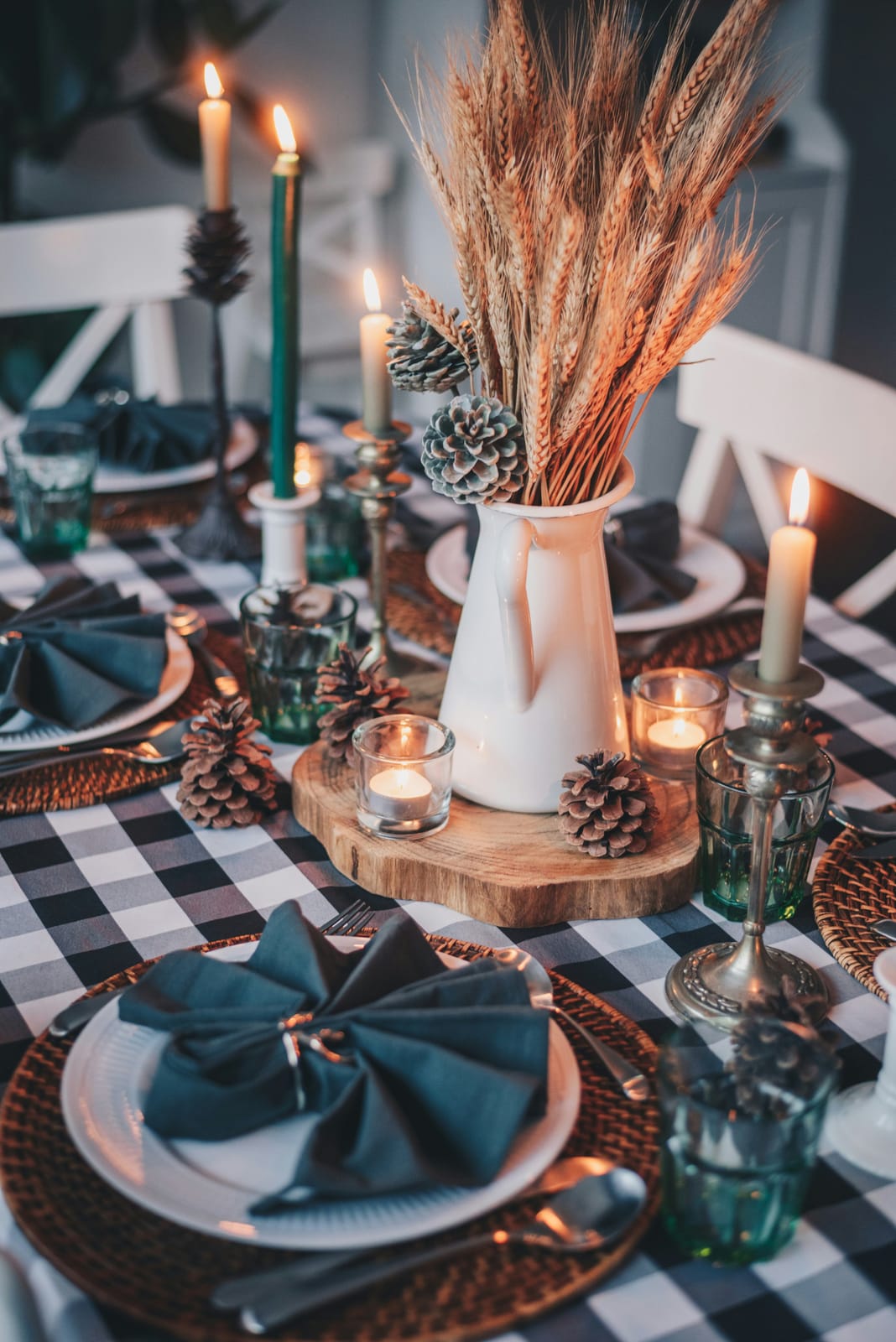 A moody fall tablescape with a black-and-white buffalo-check tablecloth, a white pitcher of dried wheat, frosted pinecones, teal napkins, and green and white taper candles