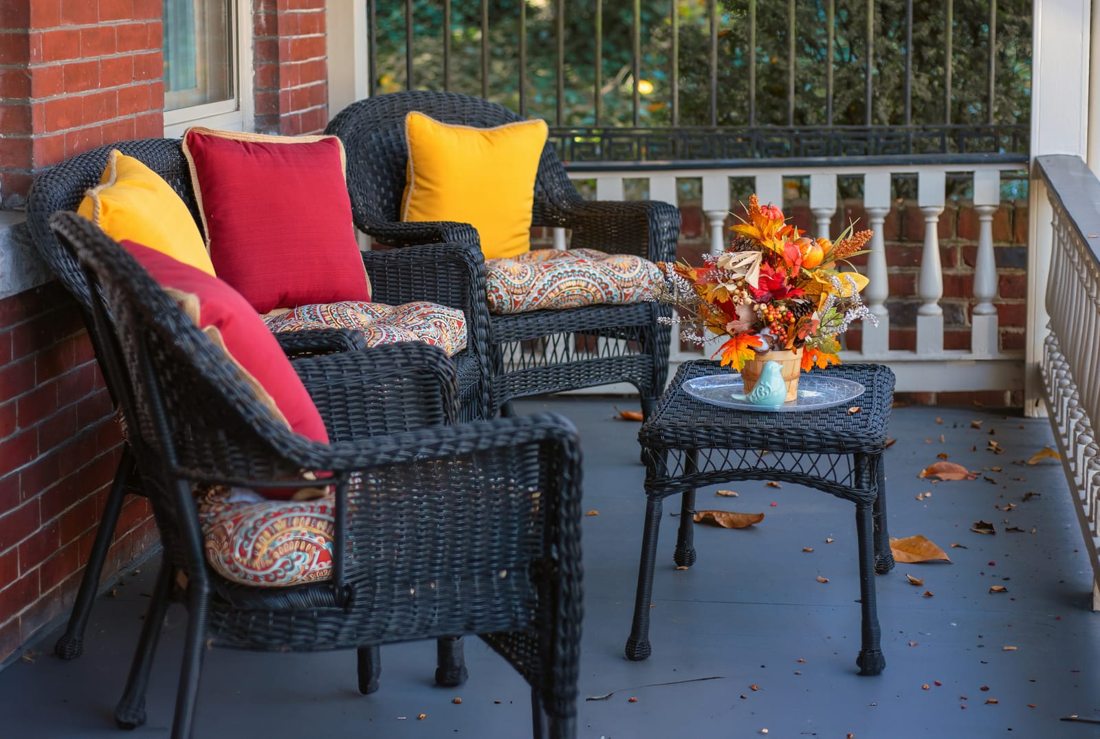 A cozy autumn porch with black wicker chairs, warm-colored pillows, and a small fall floral arrangement in soft light