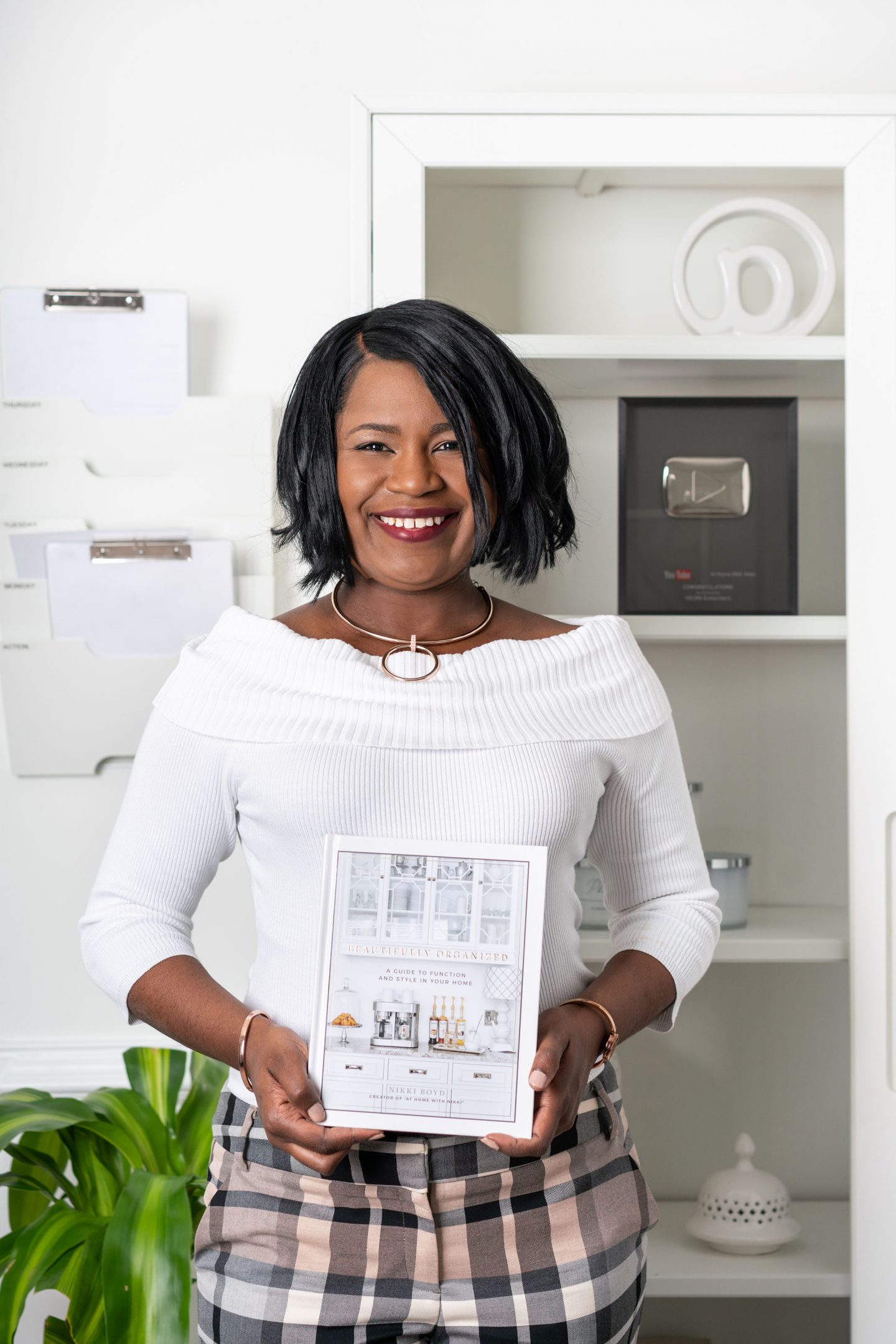 Nikki Boyd smiling in a white off-the-shoulder top, holding her book Beautifully Organized in a bright, organized home office