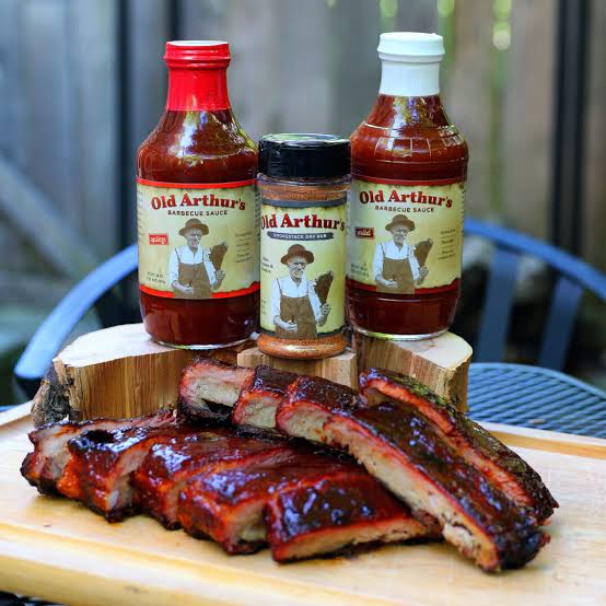 Two bottles of Old Arthur’s barbecue sauce and a dry rub shaker on wood rounds, with a sliced rack of glazed ribs