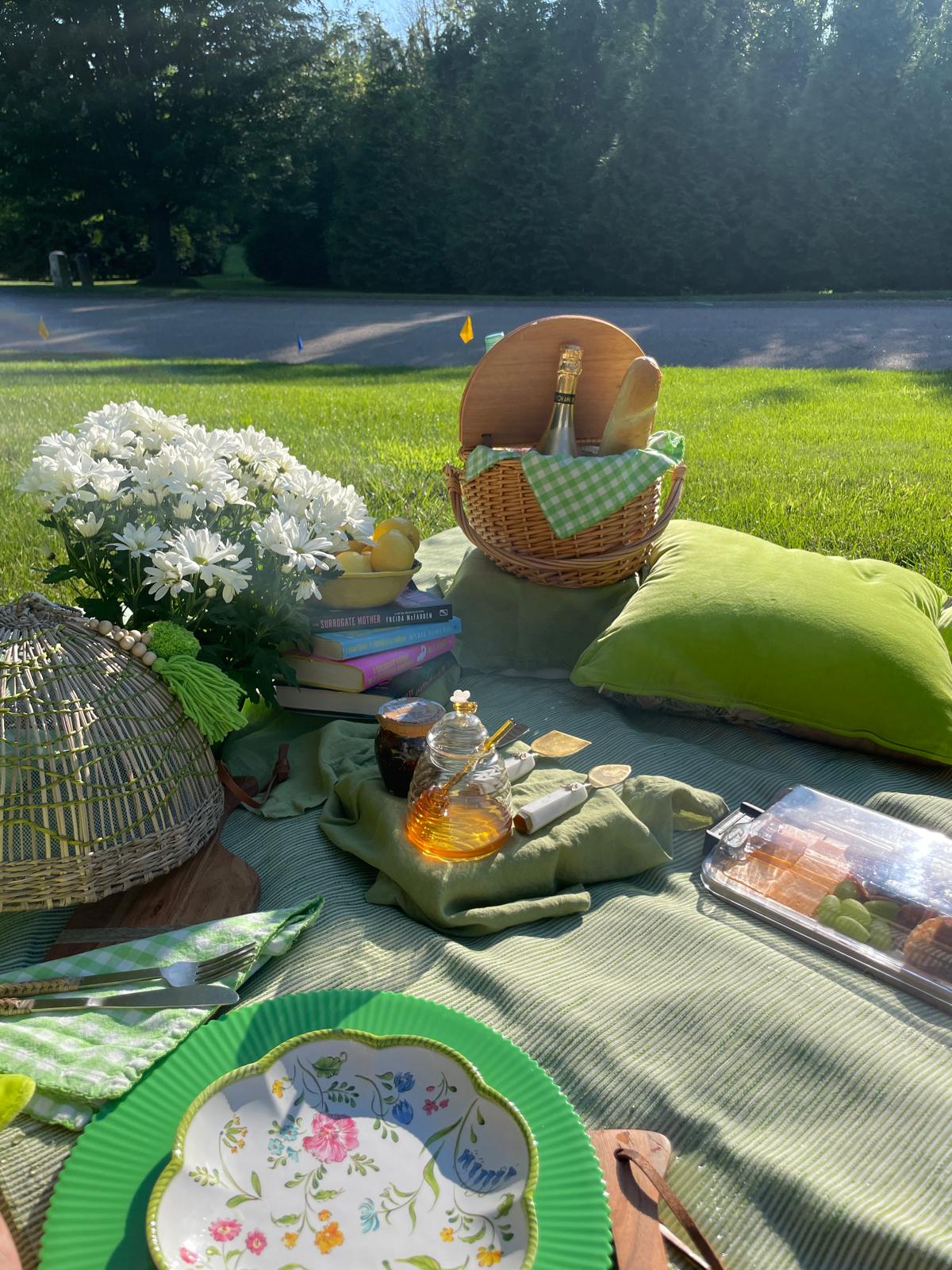 A picnic spread on a green striped blanket on a grassy lawn with daisies, a bowl of lemons, a stack of books, a wicker basket, and green velvet pillows